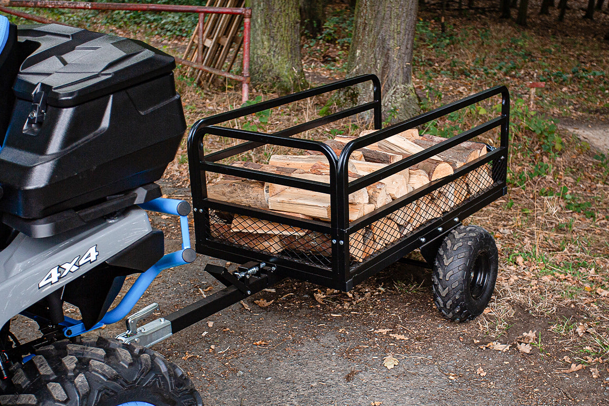 Black metal trailer attached to a 4x4 vehicle with logs inside, on a forest path.